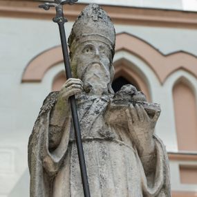 Zdjęcie nr 1: Rzeźba przedstawiająca św. Mikołaja. Święty ukazany jest w postawie stojącej, frontalnie, z krzyżem w prawej dłoni i otwartą księgą w lewej, na której ułożone są trzy kule. Twarz podłużna, okolona długą i bujną brodą, nos długi i wąski, oczy osadzone głęboko w oczodołach ze wzrokiem skierowanym na wprost; włosy długie i opadające na plecy. Święty ubrany jest w długą, przewiązaną sznurem albę, stułę, kapę zawiązaną pod szyją, infułę na głowie, zdobioną na przodzie krzyżykiem greckim, a na dłoniach ma założone rękawiczki. Krzyż na długim, metalowym drzewcu o ramionach zakończonych trójlistnie.