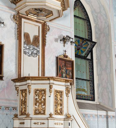 Zdjęcie nr 4: Ambona wisząca, przyścienna, z wejściem od prawej strony. Kosz na planie ośmioboku, na niskim cokole, od dołu ujęty ośmiobocznym podwieszeniem. Ścianki kosza zdobione prostokątnymi płycinami wypełnionymi stylizowaną wicią liści akantu z czteropłatkowym kwiatkiem pośrodku. Parapet w formie prostego belkowania, którego imposty wsparte są małymi hermami – główkami połączonymi z dekoracją naśladującą ornament małżowinowo-chrząstkowy. Zaplecek w kształcie stojącego prostokąta w profilowanej i złoconej ramie, w górnej partii ujęty po bokach wicią akantu ze stylizowanymi podwieszonymi kampanulami; w polu umieszczone tablice dekalogu na obłokach. Baldachim  na planie ośmioboku, z wydatnym, profilowanym gzymsem, zwieńczony kabłąkami spiętymi i podtrzymującymi krzyżyk łaciński zatknięty na kuli. Na podniebieniu baldachimu, w ośmiobocznej płycinie rzeźbiona gołębica Ducha Świętego na tle promienistej glorii złożonej z wiązek promieni o nierównej długości. Balustrada schodów zdobiona wydłużonym, stylizowanym ornamentem małżowinowo-chrząstkowym umieszczonym w płytkiej, romboidalnej płycinie. Struktura polichromowana w kolorze beżowym, profile i ornamenty złocone oraz posrebrzone.