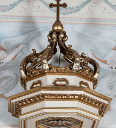 Zdjęcie nr 5: Ambona wisząca, przyścienna, z wejściem od prawej strony. Kosz na planie ośmioboku, na niskim cokole, od dołu ujęty ośmiobocznym podwieszeniem. Ścianki kosza zdobione prostokątnymi płycinami wypełnionymi stylizowaną wicią liści akantu z czteropłatkowym kwiatkiem pośrodku. Parapet w formie prostego belkowania, którego imposty wsparte są małymi hermami – główkami połączonymi z dekoracją naśladującą ornament małżowinowo-chrząstkowy. Zaplecek w kształcie stojącego prostokąta w profilowanej i złoconej ramie, w górnej partii ujęty po bokach wicią akantu ze stylizowanymi podwieszonymi kampanulami; w polu umieszczone tablice dekalogu na obłokach. Baldachim  na planie ośmioboku, z wydatnym, profilowanym gzymsem, zwieńczony kabłąkami spiętymi i podtrzymującymi krzyżyk łaciński zatknięty na kuli. Na podniebieniu baldachimu, w ośmiobocznej płycinie rzeźbiona gołębica Ducha Świętego na tle promienistej glorii złożonej z wiązek promieni o nierównej długości. Balustrada schodów zdobiona wydłużonym, stylizowanym ornamentem małżowinowo-chrząstkowym umieszczonym w płytkiej, romboidalnej płycinie. Struktura polichromowana w kolorze beżowym, profile i ornamenty złocone oraz posrebrzone.