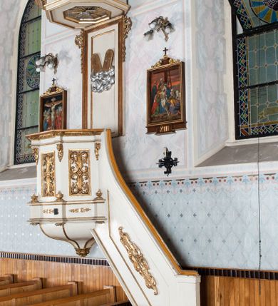 Zdjęcie nr 3: Ambona wisząca, przyścienna, z wejściem od prawej strony. Kosz na planie ośmioboku, na niskim cokole, od dołu ujęty ośmiobocznym podwieszeniem. Ścianki kosza zdobione prostokątnymi płycinami wypełnionymi stylizowaną wicią liści akantu z czteropłatkowym kwiatkiem pośrodku. Parapet w formie prostego belkowania, którego imposty wsparte są małymi hermami – główkami połączonymi z dekoracją naśladującą ornament małżowinowo-chrząstkowy. Zaplecek w kształcie stojącego prostokąta w profilowanej i złoconej ramie, w górnej partii ujęty po bokach wicią akantu ze stylizowanymi podwieszonymi kampanulami; w polu umieszczone tablice dekalogu na obłokach. Baldachim  na planie ośmioboku, z wydatnym, profilowanym gzymsem, zwieńczony kabłąkami spiętymi i podtrzymującymi krzyżyk łaciński zatknięty na kuli. Na podniebieniu baldachimu, w ośmiobocznej płycinie rzeźbiona gołębica Ducha Świętego na tle promienistej glorii złożonej z wiązek promieni o nierównej długości. Balustrada schodów zdobiona wydłużonym, stylizowanym ornamentem małżowinowo-chrząstkowym umieszczonym w płytkiej, romboidalnej płycinie. Struktura polichromowana w kolorze beżowym, profile i ornamenty złocone oraz posrebrzone.