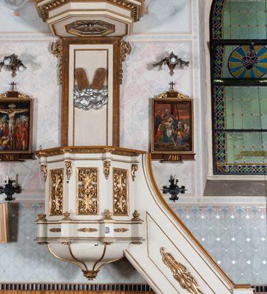 Zdjęcie nr 2: Ambona wisząca, przyścienna, z wejściem od prawej strony. Kosz na planie ośmioboku, na niskim cokole, od dołu ujęty ośmiobocznym podwieszeniem. Ścianki kosza zdobione prostokątnymi płycinami wypełnionymi stylizowaną wicią liści akantu z czteropłatkowym kwiatkiem pośrodku. Parapet w formie prostego belkowania, którego imposty wsparte są małymi hermami – główkami połączonymi z dekoracją naśladującą ornament małżowinowo-chrząstkowy. Zaplecek w kształcie stojącego prostokąta w profilowanej i złoconej ramie, w górnej partii ujęty po bokach wicią akantu ze stylizowanymi podwieszonymi kampanulami; w polu umieszczone tablice dekalogu na obłokach. Baldachim  na planie ośmioboku, z wydatnym, profilowanym gzymsem, zwieńczony kabłąkami spiętymi i podtrzymującymi krzyżyk łaciński zatknięty na kuli. Na podniebieniu baldachimu, w ośmiobocznej płycinie rzeźbiona gołębica Ducha Świętego na tle promienistej glorii złożonej z wiązek promieni o nierównej długości. Balustrada schodów zdobiona wydłużonym, stylizowanym ornamentem małżowinowo-chrząstkowym umieszczonym w płytkiej, romboidalnej płycinie. Struktura polichromowana w kolorze beżowym, profile i ornamenty złocone oraz posrebrzone.