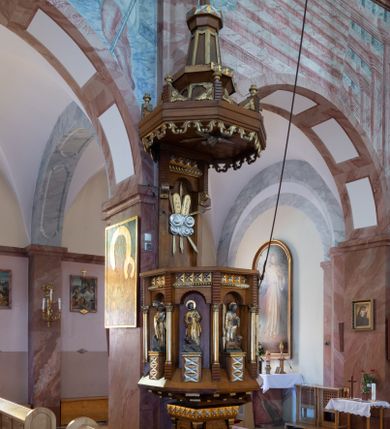 Zdjęcie nr 1: Ambona przyścienna, ustawiona na niskim filarze, dostępna od prawej strony schodami umieszczonymi przy filarze międzynawowym. Filar czworoboczny, na profilowanym cokole, naroża opięte filarkami o czworobocznych trzonach, zamknięty prostym gzymsem. Na ściankach płytkie płyciny w kształcie prostokąta o wklęsłych narożach, w złoconych obramieniach. Gzyms zdobiony rzędem niskich ostrosłupów ozdobionych rzeźbionym i złoconym greckim krzyżykiem. Filar wspiera kosz na rzucie siedmioboku, opięty na narożach kolumienkami wspierającymi belkowanie i gzyms. Ścianki kosza zdobione prostokątnymi niszami zamkniętymi łukiem półkolistym nadwieszonym. Nad niszami fryz zdobiony analogicznie jak w podstawie ambony ostrosłupami z krzyżykami greckimi, poniżej fryz z motywów rybiej łuski. We niszach, na wysokich postumentach ustawione rzeźby od lewej: kielich, św. Jan, św. Łukasz, św. Marek i św. Mateusz. Kolumny o gładkich, złoconych trzonach i kapitelach kompozytowych. Parapet w formie prostego gzymsu. Zaplecek wysoki, z płyciną zamkniętą łukiem półkolistym nadwieszonym. W polu płaskorzeźbione tablice Dekalogu umieszczone na obłokach, otoczone sześcioma wiązkami promieni o nierównej długości. Belkowanie zdobione analogicznie jak na koszu. Baldachim na rzucie ośmioboku, w formie ostrosłupa, z wydzielonym cokołem, podzielony na dwie strefy, zwieńczony umieszczonym na niskim cokole krzyżem łacińskim z odchodzącymi ze skrzyżowania ramion grubymi promieniami. Cokół baldachimu ozdobiony nad narożnikami małymi wazonikami na wysokich postumentach oraz podwieszonym lambrekinem z liści akantu. Na podniebieniu rzeźbiona Gołębica Ducha Świętego otoczona wiązkami promieni nierównej długości. Ewangeliści w niszach ukazani w pozycji stojącej, ubrani w długie i złocone szaty. Twarze szerokie, z małymi i wąskimi ustami, włosy średniej długości, ciemnobrązowe. Wokół głów złote i koliste nimby. Św. Jan zwrócony en trois quarts w lewo, z otwartą księgą Pisma Świętego w lewej dłoni, w której napis: A SŁOWO BYŁO U BOGA / I BOGIEM BYŁO SŁOWO. Św. Łukasz ukazany en face, z głową zwróconą w lewo. Św. Marek ukazany en face, z głową zwróconą w prawo. Po jego prawej stronie kolumna. Św. Mateusz ukazany frontalnie z otwartą księgą w dłoni, w tle, po jego prawej stronie drzewo. Z prawej strony jednobiegowe schody z balustradą zakończoną słupkiem w formie filaru, z arkadami zamkniętymi łukiem półkolistym. Struktura malowana w kolorze jasnobrązowym, profile i ornamenty złocone oraz srebrzone.