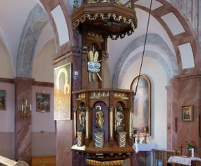 Zdjęcie nr 1: Ambona przyścienna, ustawiona na niskim filarze, dostępna od prawej strony schodami umieszczonymi przy filarze międzynawowym. Filar czworoboczny, na profilowanym cokole, naroża opięte filarkami o czworobocznych trzonach, zamknięty prostym gzymsem. Na ściankach płytkie płyciny w kształcie prostokąta o wklęsłych narożach, w złoconych obramieniach. Gzyms zdobiony rzędem niskich ostrosłupów ozdobionych rzeźbionym i złoconym greckim krzyżykiem. Filar wspiera kosz na rzucie siedmioboku, opięty na narożach kolumienkami wspierającymi belkowanie i gzyms. Ścianki kosza zdobione prostokątnymi niszami zamkniętymi łukiem półkolistym nadwieszonym. Nad niszami fryz zdobiony analogicznie jak w podstawie ambony ostrosłupami z krzyżykami greckimi, poniżej fryz z motywów rybiej łuski. We niszach, na wysokich postumentach ustawione rzeźby od lewej: kielich, św. Jan, św. Łukasz, św. Marek i św. Mateusz. Kolumny o gładkich, złoconych trzonach i kapitelach kompozytowych. Parapet w formie prostego gzymsu. Zaplecek wysoki, z płyciną zamkniętą łukiem półkolistym nadwieszonym. W polu płaskorzeźbione tablice Dekalogu umieszczone na obłokach, otoczone sześcioma wiązkami promieni o nierównej długości. Belkowanie zdobione analogicznie jak na koszu. Baldachim na rzucie ośmioboku, w formie ostrosłupa, z wydzielonym cokołem, podzielony na dwie strefy, zwieńczony umieszczonym na niskim cokole krzyżem łacińskim z odchodzącymi ze skrzyżowania ramion grubymi promieniami. Cokół baldachimu ozdobiony nad narożnikami małymi wazonikami na wysokich postumentach oraz podwieszonym lambrekinem z liści akantu. Na podniebieniu rzeźbiona Gołębica Ducha Świętego otoczona wiązkami promieni nierównej długości. Ewangeliści w niszach ukazani w pozycji stojącej, ubrani w długie i złocone szaty. Twarze szerokie, z małymi i wąskimi ustami, włosy średniej długości, ciemnobrązowe. Wokół głów złote i koliste nimby. Św. Jan zwrócony en trois quarts w lewo, z otwartą księgą Pisma Świętego w lewej dłoni, w której napis: A SŁOWO BYŁO U BOGA / I BOGIEM BYŁO SŁOWO. Św. Łukasz ukazany en face, z głową zwróconą w lewo. Św. Marek ukazany en face, z głową zwróconą w prawo. Po jego prawej stronie kolumna. Św. Mateusz ukazany frontalnie z otwartą księgą w dłoni, w tle, po jego prawej stronie drzewo. Z prawej strony jednobiegowe schody z balustradą zakończoną słupkiem w formie filaru, z arkadami zamkniętymi łukiem półkolistym. Struktura malowana w kolorze jasnobrązowym, profile i ornamenty złocone oraz srebrzone.