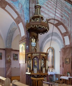 Zdjęcie nr 1: Ambona przyścienna, ustawiona na niskim filarze, dostępna od prawej strony schodami umieszczonymi przy filarze międzynawowym. Filar czworoboczny, na profilowanym cokole, naroża opięte filarkami o czworobocznych trzonach, zamknięty prostym gzymsem. Na ściankach płytkie płyciny w kształcie prostokąta o wklęsłych narożach, w złoconych obramieniach. Gzyms zdobiony rzędem niskich ostrosłupów ozdobionych rzeźbionym i złoconym greckim krzyżykiem. Filar wspiera kosz na rzucie siedmioboku, opięty na narożach kolumienkami wspierającymi belkowanie i gzyms. Ścianki kosza zdobione prostokątnymi niszami zamkniętymi łukiem półkolistym nadwieszonym. Nad niszami fryz zdobiony analogicznie jak w podstawie ambony ostrosłupami z krzyżykami greckimi, poniżej fryz z motywów rybiej łuski. We niszach, na wysokich postumentach ustawione rzeźby od lewej: kielich, św. Jan, św. Łukasz, św. Marek i św. Mateusz. Kolumny o gładkich, złoconych trzonach i kapitelach kompozytowych. Parapet w formie prostego gzymsu. Zaplecek wysoki, z płyciną zamkniętą łukiem półkolistym nadwieszonym. W polu płaskorzeźbione tablice Dekalogu umieszczone na obłokach, otoczone sześcioma wiązkami promieni o nierównej długości. Belkowanie zdobione analogicznie jak na koszu. Baldachim na rzucie ośmioboku, w formie ostrosłupa, z wydzielonym cokołem, podzielony na dwie strefy, zwieńczony umieszczonym na niskim cokole krzyżem łacińskim z odchodzącymi ze skrzyżowania ramion grubymi promieniami. Cokół baldachimu ozdobiony nad narożnikami małymi wazonikami na wysokich postumentach oraz podwieszonym lambrekinem z liści akantu. Na podniebieniu rzeźbiona Gołębica Ducha Świętego otoczona wiązkami promieni nierównej długości. Ewangeliści w niszach ukazani w pozycji stojącej, ubrani w długie i złocone szaty. Twarze szerokie, z małymi i wąskimi ustami, włosy średniej długości, ciemnobrązowe. Wokół głów złote i koliste nimby. Św. Jan zwrócony en trois quarts w lewo, z otwartą księgą Pisma Świętego w lewej dłoni, w której napis: A SŁOWO BYŁO U BOGA / I BOGIEM BYŁO SŁOWO. Św. Łukasz ukazany en face, z głową zwróconą w lewo. Św. Marek ukazany en face, z głową zwróconą w prawo. Po jego prawej stronie kolumna. Św. Mateusz ukazany frontalnie z otwartą księgą w dłoni, w tle, po jego prawej stronie drzewo. Z prawej strony jednobiegowe schody z balustradą zakończoną słupkiem w formie filaru, z arkadami zamkniętymi łukiem półkolistym. Struktura malowana w kolorze jasnobrązowym, profile i ornamenty złocone oraz srebrzone.