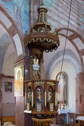 Zdjęcie nr 1: Ambona przyścienna, ustawiona na niskim filarze, dostępna od prawej strony schodami umieszczonymi przy filarze międzynawowym. Filar czworoboczny, na profilowanym cokole, naroża opięte filarkami o czworobocznych trzonach, zamknięty prostym gzymsem. Na ściankach płytkie płyciny w kształcie prostokąta o wklęsłych narożach, w złoconych obramieniach. Gzyms zdobiony rzędem niskich ostrosłupów ozdobionych rzeźbionym i złoconym greckim krzyżykiem. Filar wspiera kosz na rzucie siedmioboku, opięty na narożach kolumienkami wspierającymi belkowanie i gzyms. Ścianki kosza zdobione prostokątnymi niszami zamkniętymi łukiem półkolistym nadwieszonym. Nad niszami fryz zdobiony analogicznie jak w podstawie ambony ostrosłupami z krzyżykami greckimi, poniżej fryz z motywów rybiej łuski. We niszach, na wysokich postumentach ustawione rzeźby od lewej: kielich, św. Jan, św. Łukasz, św. Marek i św. Mateusz. Kolumny o gładkich, złoconych trzonach i kapitelach kompozytowych. Parapet w formie prostego gzymsu. Zaplecek wysoki, z płyciną zamkniętą łukiem półkolistym nadwieszonym. W polu płaskorzeźbione tablice Dekalogu umieszczone na obłokach, otoczone sześcioma wiązkami promieni o nierównej długości. Belkowanie zdobione analogicznie jak na koszu. Baldachim na rzucie ośmioboku, w formie ostrosłupa, z wydzielonym cokołem, podzielony na dwie strefy, zwieńczony umieszczonym na niskim cokole krzyżem łacińskim z odchodzącymi ze skrzyżowania ramion grubymi promieniami. Cokół baldachimu ozdobiony nad narożnikami małymi wazonikami na wysokich postumentach oraz podwieszonym lambrekinem z liści akantu. Na podniebieniu rzeźbiona Gołębica Ducha Świętego otoczona wiązkami promieni nierównej długości. Ewangeliści w niszach ukazani w pozycji stojącej, ubrani w długie i złocone szaty. Twarze szerokie, z małymi i wąskimi ustami, włosy średniej długości, ciemnobrązowe. Wokół głów złote i koliste nimby. Św. Jan zwrócony en trois quarts w lewo, z otwartą księgą Pisma Świętego w lewej dłoni, w której napis: A SŁOWO BYŁO U BOGA / I BOGIEM BYŁO SŁOWO. Św. Łukasz ukazany en face, z głową zwróconą w lewo. Św. Marek ukazany en face, z głową zwróconą w prawo. Po jego prawej stronie kolumna. Św. Mateusz ukazany frontalnie z otwartą księgą w dłoni, w tle, po jego prawej stronie drzewo. Z prawej strony jednobiegowe schody z balustradą zakończoną słupkiem w formie filaru, z arkadami zamkniętymi łukiem półkolistym. Struktura malowana w kolorze jasnobrązowym, profile i ornamenty złocone oraz srebrzone.