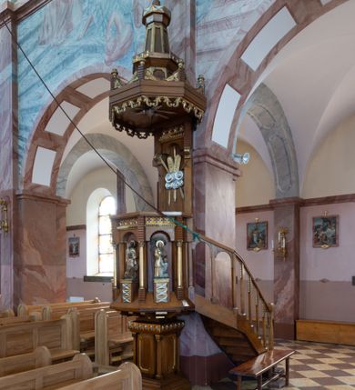 Zdjęcie nr 2: Ambona przyścienna, ustawiona na niskim filarze, dostępna od prawej strony schodami umieszczonymi przy filarze międzynawowym. Filar czworoboczny, na profilowanym cokole, naroża opięte filarkami o czworobocznych trzonach, zamknięty prostym gzymsem. Na ściankach płytkie płyciny w kształcie prostokąta o wklęsłych narożach, w złoconych obramieniach. Gzyms zdobiony rzędem niskich ostrosłupów ozdobionych rzeźbionym i złoconym greckim krzyżykiem. Filar wspiera kosz na rzucie siedmioboku, opięty na narożach kolumienkami wspierającymi belkowanie i gzyms. Ścianki kosza zdobione prostokątnymi niszami zamkniętymi łukiem półkolistym nadwieszonym. Nad niszami fryz zdobiony analogicznie jak w podstawie ambony ostrosłupami z krzyżykami greckimi, poniżej fryz z motywów rybiej łuski. We niszach, na wysokich postumentach ustawione rzeźby od lewej: kielich, św. Jan, św. Łukasz, św. Marek i św. Mateusz. Kolumny o gładkich, złoconych trzonach i kapitelach kompozytowych. Parapet w formie prostego gzymsu. Zaplecek wysoki, z płyciną zamkniętą łukiem półkolistym nadwieszonym. W polu płaskorzeźbione tablice Dekalogu umieszczone na obłokach, otoczone sześcioma wiązkami promieni o nierównej długości. Belkowanie zdobione analogicznie jak na koszu. Baldachim na rzucie ośmioboku, w formie ostrosłupa, z wydzielonym cokołem, podzielony na dwie strefy, zwieńczony umieszczonym na niskim cokole krzyżem łacińskim z odchodzącymi ze skrzyżowania ramion grubymi promieniami. Cokół baldachimu ozdobiony nad narożnikami małymi wazonikami na wysokich postumentach oraz podwieszonym lambrekinem z liści akantu. Na podniebieniu rzeźbiona Gołębica Ducha Świętego otoczona wiązkami promieni nierównej długości. Ewangeliści w niszach ukazani w pozycji stojącej, ubrani w długie i złocone szaty. Twarze szerokie, z małymi i wąskimi ustami, włosy średniej długości, ciemnobrązowe. Wokół głów złote i koliste nimby. Św. Jan zwrócony en trois quarts w lewo, z otwartą księgą Pisma Świętego w lewej dłoni, w której napis: A SŁOWO BYŁO U BOGA / I BOGIEM BYŁO SŁOWO. Św. Łukasz ukazany en face, z głową zwróconą w lewo. Św. Marek ukazany en face, z głową zwróconą w prawo. Po jego prawej stronie kolumna. Św. Mateusz ukazany frontalnie z otwartą księgą w dłoni, w tle, po jego prawej stronie drzewo. Z prawej strony jednobiegowe schody z balustradą zakończoną słupkiem w formie filaru, z arkadami zamkniętymi łukiem półkolistym. Struktura malowana w kolorze jasnobrązowym, profile i ornamenty złocone oraz srebrzone.