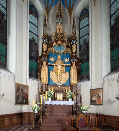 Zdjęcie nr 11: Nastawa architektoniczna, jednoosiowa, jednokondygnacyjna, ze zwieńczeniem, ustawiona na wysokim cokole. Ołtarz ustawiony na pięciostopniowej kamiennej podstawie; granitowy, prostopadłościenny, z monogramem IHS na froncie, mensa granitowa. Na nim drewniane tabernakulum w formie prostopadłościennej skrzynki, flankowane parą kolumienek, zamknięte od góry profilowanym gzymsem. Na ściance frontowej wysunięte do przodu metalowe drzwiczki w oprawie architektonicznej na niskim cokole, zamknięte od góry prostym gzymsem. Na drzwiczkach kielich z hostią, w płycinie o kształcie prostokąta stojącego zamkniętego łukiem trójlistnym ostrym. 
Nastawa flankowana dwiema dwukondygnacyjnymi wieżyczkami na planie ośmioboku z figurami św. Jana Ewangelisty i św. Piotra w pierwszej kondygnacji oraz figurami aniołów w drugiej. Figury apostołów ustawione są na ośmiobocznych wspornikach zdobionych od dołu arkadowaniem z kwiatonami, podtrzymywanych przez ośmioboczne kolumienki, flankujące kamienny ołtarz. Nad figurami ośmioboczne baldachimy zdobione od dołu arkadowaniem z kwiatonami, stanowiące podstawę dla figur aniołów, które zwieńczone są analogicznymi ośmiobocznymi baldachimami i rozbudowanymi pinaklami. W polu środkowym rzeźbiona Trójca Święta ukazana jako Tron łaski pośród obłoków i uskrzydlonych główek anielskich. Nad nią duży, ośmioboczny baldachim, zdobiony od dołu arkadowaniem, zakończonym kwiatonami, dekorowany maswerkami, wimpergami i kwiatonami, naprzemiennie z pinaklami, który stanowi podstawę wieżyczki wieńczącej ołtarz. W jej polu ustawiona figura Matki Boskiej, nad nią ośmioboczny baldachim zdobiony od dołu arkadowaniem i kwiatonami, zakończony dużym pinaklem dekorowanym żabkami. Nastawa flankowana uszami w formie przypór dekorowanych maswerkiem i wąskimi kolumienkami z pinaklami. 
Struktura politurowana w kolorze ciemnobrązowym, detale i ornamentyka złocone, tło pola głównego oraz podniebienia baldachimów błękitne.