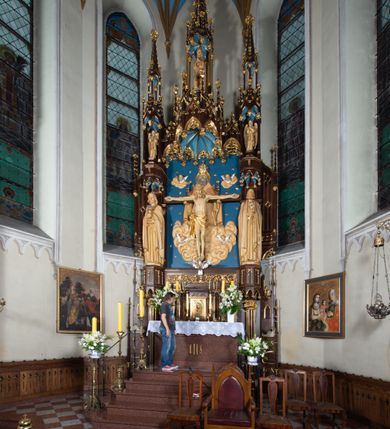 Zdjęcie nr 16: Nastawa architektoniczna, jednoosiowa, jednokondygnacyjna, ze zwieńczeniem, ustawiona na wysokim cokole. Ołtarz ustawiony na pięciostopniowej kamiennej podstawie; granitowy, prostopadłościenny, z monogramem IHS na froncie, mensa granitowa. Na nim drewniane tabernakulum w formie prostopadłościennej skrzynki, flankowane parą kolumienek, zamknięte od góry profilowanym gzymsem. Na ściance frontowej wysunięte do przodu metalowe drzwiczki w oprawie architektonicznej na niskim cokole, zamknięte od góry prostym gzymsem. Na drzwiczkach kielich z hostią, w płycinie o kształcie prostokąta stojącego zamkniętego łukiem trójlistnym ostrym. 
Nastawa flankowana dwiema dwukondygnacyjnymi wieżyczkami na planie ośmioboku z figurami św. Jana Ewangelisty i św. Piotra w pierwszej kondygnacji oraz figurami aniołów w drugiej. Figury apostołów ustawione są na ośmiobocznych wspornikach zdobionych od dołu arkadowaniem z kwiatonami, podtrzymywanych przez ośmioboczne kolumienki, flankujące kamienny ołtarz. Nad figurami ośmioboczne baldachimy zdobione od dołu arkadowaniem z kwiatonami, stanowiące podstawę dla figur aniołów, które zwieńczone są analogicznymi ośmiobocznymi baldachimami i rozbudowanymi pinaklami. W polu środkowym rzeźbiona Trójca Święta ukazana jako Tron łaski pośród obłoków i uskrzydlonych główek anielskich. Nad nią duży, ośmioboczny baldachim, zdobiony od dołu arkadowaniem, zakończonym kwiatonami, dekorowany maswerkami, wimpergami i kwiatonami, naprzemiennie z pinaklami, który stanowi podstawę wieżyczki wieńczącej ołtarz. W jej polu ustawiona figura Matki Boskiej, nad nią ośmioboczny baldachim zdobiony od dołu arkadowaniem i kwiatonami, zakończony dużym pinaklem dekorowanym żabkami. Nastawa flankowana uszami w formie przypór dekorowanych maswerkiem i wąskimi kolumienkami z pinaklami. 
Struktura politurowana w kolorze ciemnobrązowym, detale i ornamentyka złocone, tło pola głównego oraz podniebienia baldachimów błękitne.