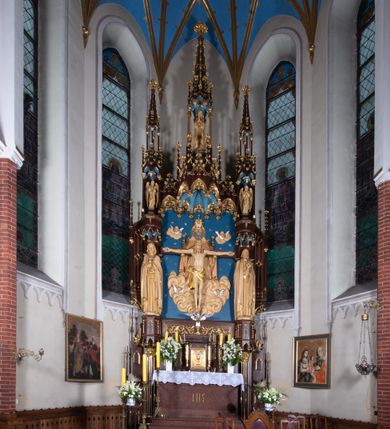 Zdjęcie nr 2: Nastawa architektoniczna, jednoosiowa, jednokondygnacyjna, ze zwieńczeniem, ustawiona na wysokim cokole. Ołtarz ustawiony na pięciostopniowej kamiennej podstawie; granitowy, prostopadłościenny, z monogramem IHS na froncie, mensa granitowa. Na nim drewniane tabernakulum w formie prostopadłościennej skrzynki, flankowane parą kolumienek, zamknięte od góry profilowanym gzymsem. Na ściance frontowej wysunięte do przodu metalowe drzwiczki w oprawie architektonicznej na niskim cokole, zamknięte od góry prostym gzymsem. Na drzwiczkach kielich z hostią, w płycinie o kształcie prostokąta stojącego zamkniętego łukiem trójlistnym ostrym. 
Nastawa flankowana dwiema dwukondygnacyjnymi wieżyczkami na planie ośmioboku z figurami św. Jana Ewangelisty i św. Piotra w pierwszej kondygnacji oraz figurami aniołów w drugiej. Figury apostołów ustawione są na ośmiobocznych wspornikach zdobionych od dołu arkadowaniem z kwiatonami, podtrzymywanych przez ośmioboczne kolumienki, flankujące kamienny ołtarz. Nad figurami ośmioboczne baldachimy zdobione od dołu arkadowaniem z kwiatonami, stanowiące podstawę dla figur aniołów, które zwieńczone są analogicznymi ośmiobocznymi baldachimami i rozbudowanymi pinaklami. W polu środkowym rzeźbiona Trójca Święta ukazana jako Tron łaski pośród obłoków i uskrzydlonych główek anielskich. Nad nią duży, ośmioboczny baldachim, zdobiony od dołu arkadowaniem, zakończonym kwiatonami, dekorowany maswerkami, wimpergami i kwiatonami, naprzemiennie z pinaklami, który stanowi podstawę wieżyczki wieńczącej ołtarz. W jej polu ustawiona figura Matki Boskiej, nad nią ośmioboczny baldachim zdobiony od dołu arkadowaniem i kwiatonami, zakończony dużym pinaklem dekorowanym żabkami. Nastawa flankowana uszami w formie przypór dekorowanych maswerkiem i wąskimi kolumienkami z pinaklami. 
Struktura politurowana w kolorze ciemnobrązowym, detale i ornamentyka złocone, tło pola głównego oraz podniebienia baldachimów błękitne.
