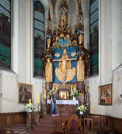 Zdjęcie nr 15: Nastawa architektoniczna, jednoosiowa, jednokondygnacyjna, ze zwieńczeniem, ustawiona na wysokim cokole. Ołtarz ustawiony na pięciostopniowej kamiennej podstawie; granitowy, prostopadłościenny, z monogramem IHS na froncie, mensa granitowa. Na nim drewniane tabernakulum w formie prostopadłościennej skrzynki, flankowane parą kolumienek, zamknięte od góry profilowanym gzymsem. Na ściance frontowej wysunięte do przodu metalowe drzwiczki w oprawie architektonicznej na niskim cokole, zamknięte od góry prostym gzymsem. Na drzwiczkach kielich z hostią, w płycinie o kształcie prostokąta stojącego zamkniętego łukiem trójlistnym ostrym. 
Nastawa flankowana dwiema dwukondygnacyjnymi wieżyczkami na planie ośmioboku z figurami św. Jana Ewangelisty i św. Piotra w pierwszej kondygnacji oraz figurami aniołów w drugiej. Figury apostołów ustawione są na ośmiobocznych wspornikach zdobionych od dołu arkadowaniem z kwiatonami, podtrzymywanych przez ośmioboczne kolumienki, flankujące kamienny ołtarz. Nad figurami ośmioboczne baldachimy zdobione od dołu arkadowaniem z kwiatonami, stanowiące podstawę dla figur aniołów, które zwieńczone są analogicznymi ośmiobocznymi baldachimami i rozbudowanymi pinaklami. W polu środkowym rzeźbiona Trójca Święta ukazana jako Tron łaski pośród obłoków i uskrzydlonych główek anielskich. Nad nią duży, ośmioboczny baldachim, zdobiony od dołu arkadowaniem, zakończonym kwiatonami, dekorowany maswerkami, wimpergami i kwiatonami, naprzemiennie z pinaklami, który stanowi podstawę wieżyczki wieńczącej ołtarz. W jej polu ustawiona figura Matki Boskiej, nad nią ośmioboczny baldachim zdobiony od dołu arkadowaniem i kwiatonami, zakończony dużym pinaklem dekorowanym żabkami. Nastawa flankowana uszami w formie przypór dekorowanych maswerkiem i wąskimi kolumienkami z pinaklami. 
Struktura politurowana w kolorze ciemnobrązowym, detale i ornamentyka złocone, tło pola głównego oraz podniebienia baldachimów błękitne.