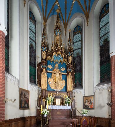 Zdjęcie nr 10: Nastawa architektoniczna, jednoosiowa, jednokondygnacyjna, ze zwieńczeniem, ustawiona na wysokim cokole. Ołtarz ustawiony na pięciostopniowej kamiennej podstawie; granitowy, prostopadłościenny, z monogramem IHS na froncie, mensa granitowa. Na nim drewniane tabernakulum w formie prostopadłościennej skrzynki, flankowane parą kolumienek, zamknięte od góry profilowanym gzymsem. Na ściance frontowej wysunięte do przodu metalowe drzwiczki w oprawie architektonicznej na niskim cokole, zamknięte od góry prostym gzymsem. Na drzwiczkach kielich z hostią, w płycinie o kształcie prostokąta stojącego zamkniętego łukiem trójlistnym ostrym. 
Nastawa flankowana dwiema dwukondygnacyjnymi wieżyczkami na planie ośmioboku z figurami św. Jana Ewangelisty i św. Piotra w pierwszej kondygnacji oraz figurami aniołów w drugiej. Figury apostołów ustawione są na ośmiobocznych wspornikach zdobionych od dołu arkadowaniem z kwiatonami, podtrzymywanych przez ośmioboczne kolumienki, flankujące kamienny ołtarz. Nad figurami ośmioboczne baldachimy zdobione od dołu arkadowaniem z kwiatonami, stanowiące podstawę dla figur aniołów, które zwieńczone są analogicznymi ośmiobocznymi baldachimami i rozbudowanymi pinaklami. W polu środkowym rzeźbiona Trójca Święta ukazana jako Tron łaski pośród obłoków i uskrzydlonych główek anielskich. Nad nią duży, ośmioboczny baldachim, zdobiony od dołu arkadowaniem, zakończonym kwiatonami, dekorowany maswerkami, wimpergami i kwiatonami, naprzemiennie z pinaklami, który stanowi podstawę wieżyczki wieńczącej ołtarz. W jej polu ustawiona figura Matki Boskiej, nad nią ośmioboczny baldachim zdobiony od dołu arkadowaniem i kwiatonami, zakończony dużym pinaklem dekorowanym żabkami. Nastawa flankowana uszami w formie przypór dekorowanych maswerkiem i wąskimi kolumienkami z pinaklami. 
Struktura politurowana w kolorze ciemnobrązowym, detale i ornamentyka złocone, tło pola głównego oraz podniebienia baldachimów błękitne.