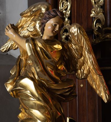 Zdjęcie nr 1: Rzeźby drążone z tyłu, klęczące na obłokach, ustawione na prostokątnych postumentach o ściętych narożach z przodu. Figury zwrócone do wewnątrz, z rękoma złożonymi w geście modlitwy, skierowanymi w przeciwną stronę. Twarze mają pociągłe, o pełnych policzkach, migdałowatych oczach, małych nosach i rozchylonych ustach. Włosy mają krótkie, brązowe, układające się w fale. Anioły ubrane są w luźne tuniki o szerokich rękawach, przepasane w talii oraz obfite płaszcze zarzucone na jedno ramię i otaczające postacie wokół nóg. Skrzydła mają rozłożone. Polichromia ciała naturalistyczna, szaty i skrzydła złocone.
