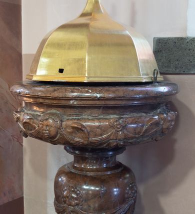 Zdjęcie nr 1: Chrzcielnica kielichowa, ustawiona na dużej podstawie na rzucie ściętego koła oraz na prostopadłościennym cokole na rzucie kwadratu. Trzon wklęsło-wypukły z uskokiem u spodu; nodus gruszkowaty, dekorowany czterema lwimi głowami trzymającymi w paszczach festony z podwieszonej tkaniny. Czara w formie bardzo spłaszczonej półkuli z wywiniętą i profilowaną krawędzią; dekorowana uskrzydlonymi główkami anielskimi na przemian z kokardami z podwieszonymi festonami z tkaniny. Pokrywa w formie baldachimu na rzucie sześcioboku, wypiętrzona, zwieńczona krzyżem.