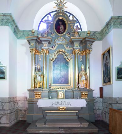 Zdjęcie nr 1: Ołtarz architektoniczny, jednoosiowy, z wydzieloną częścią cokołową i predellą, jednokondygnacyjny, ze zwieńczeniem, dostępny po dwóch kamiennych stopniach. Mensa drewniana z kamiennym portatylem, wsparta na tumbie z neorokokowym kartuszem na froncie. Tabernakulum w formie prostopadłościennej skrzynki, flankowanej z przodu płaskorzeźbionymi kolumienkami, nad nimi rozety; górna krawędź obwiedziona profilowanym gzymsem. Drzwiczki w kształcie formie stojącego prostokąta zamkniętego łukiem półkolistym nadwieszonym, ujęte ramą, z płaskorzeźbionym przedstawieniem kielicha i hostii w polu. Nastawa ujęta dwiema parami kolumn umieszczonych na tle pilastrów podtrzymujących pełne belkowanie, przerwane nad polem środkowym, występujące ponad kolumnami; skrajna para kolumn ustawiona ukośnie względem retabulum. W polu środkowym nisza w kształcie stojącego prostokąta zamkniętego łukiem wklęsło-wypukłym nadwieszonym, ujęta podwójną profilowaną ramą, w niszy obraz Trójcą Święta. Ponad obrazem, na osin neorokokowy kartusz z koroną i inicjałami: KK, otoczony czterema uskrzydlonymi główkami anielskimi. Pomiędzy kolumnami figury św. Wojciecha i św. Stanisława ustawione na konsolach. Zwieńczenie w kształcie aedikuli, ujęte parą łukowato wygiętych pilastrów oraz spływami wolutowymi, na gzymsie koronującym z płaskorzeźbionym Okiem Opatrzności w glorii promienistej oraz z parą wazonów po bokach. W polu owalny obraz z przedstawieniem Ecce Homo ujęty profilowaną ramą. W zwieńczeniu, w osiach kolumn para puttów i para wazonów. Trzony pilastrów i przyłucza w polu głównym dekorowane ornamentem neorokokowym; fryz zdobiony rozetami. Struktura polichromowana w kolorze seledynowym; kolumny, ornamenty i profile złocone.