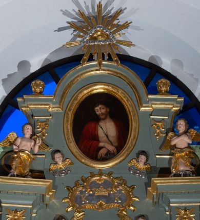 Zdjęcie nr 2: Ołtarz architektoniczny, jednoosiowy, z wydzieloną częścią cokołową i predellą, jednokondygnacyjny, ze zwieńczeniem, dostępny po dwóch kamiennych stopniach. Mensa drewniana z kamiennym portatylem, wsparta na tumbie z neorokokowym kartuszem na froncie. Tabernakulum w formie prostopadłościennej skrzynki, flankowanej z przodu płaskorzeźbionymi kolumienkami, nad nimi rozety; górna krawędź obwiedziona profilowanym gzymsem. Drzwiczki w kształcie formie stojącego prostokąta zamkniętego łukiem półkolistym nadwieszonym, ujęte ramą, z płaskorzeźbionym przedstawieniem kielicha i hostii w polu. Nastawa ujęta dwiema parami kolumn umieszczonych na tle pilastrów podtrzymujących pełne belkowanie, przerwane nad polem środkowym, występujące ponad kolumnami; skrajna para kolumn ustawiona ukośnie względem retabulum. W polu środkowym nisza w kształcie stojącego prostokąta zamkniętego łukiem wklęsło-wypukłym nadwieszonym, ujęta podwójną profilowaną ramą, w niszy obraz Trójcą Święta. Ponad obrazem, na osin neorokokowy kartusz z koroną i inicjałami: KK, otoczony czterema uskrzydlonymi główkami anielskimi. Pomiędzy kolumnami figury św. Wojciecha i św. Stanisława ustawione na konsolach. Zwieńczenie w kształcie aedikuli, ujęte parą łukowato wygiętych pilastrów oraz spływami wolutowymi, na gzymsie koronującym z płaskorzeźbionym Okiem Opatrzności w glorii promienistej oraz z parą wazonów po bokach. W polu owalny obraz z przedstawieniem Ecce Homo ujęty profilowaną ramą. W zwieńczeniu, w osiach kolumn para puttów i para wazonów. Trzony pilastrów i przyłucza w polu głównym dekorowane ornamentem neorokokowym; fryz zdobiony rozetami. Struktura polichromowana w kolorze seledynowym; kolumny, ornamenty i profile złocone.