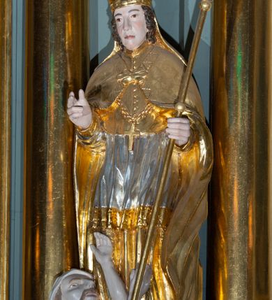 Zdjęcie nr 3: Rzeźba pełnoplastyczna, przyścienna, z tyłu drążona, ustawiona na niskim, prostopadłościennym postumencie. Święty ukazany en face, w kontrapoście, obie ręce ugięte w łokciach, wyciągnięte przed siebie, w lewej trzyma pastorał, prawa dłoń ułożona w geście błogosławieństwa. Twarz pociągła, o płytko rzeźbionych rysach i dużym, zaostrzonym nosie, okolona krótkimi falowanymi włosami. Ubrany w długą albę, komżę z koronką przy brzegach, mucet zapinany na guziki oraz kapę spiętą na piersi i opadającą na ramiona i plecy, z fragmentem tkaniny zawiniętym przy lewym boku; na piersi pektorał,  na głowie infuła. Przy prawej nodze Piotrowin, ukazany w popiersiu, z profilu, z rękoma uniesionymi ku górze oraz głową okrytą całunem. Figura polichromowana naturalistycznie w odsłoniętych partiach ciała, szaty i atrybuty złocone, komża srebrzona.