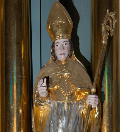 Zdjęcie nr 1: Rzeźba pełnoplastyczna, przyścienna, z tyłu drążona, ustawiona na niskim, prostopadłościennym postumencie. Święty ukazany en face, w kontrapoście, obie ręce ugięte w łokciach, wyciągnięte przed siebie, w lewej trzyma pastorał, prawa dłoń ułożona w geście błogosławieństwa. Twarz pociągła, o płytko rzeźbionych rysach i dużym, zaostrzonym nosie, okolona krótkimi falowanymi włosami. Ubrany w długą albę, komżę z koronką przy brzegach, mucet zapinany na guziki oraz kapę spiętą na piersi i opadającą na ramiona i plecy, z fragmentem tkaniny zawiniętym przy lewym boku; na piersi pektorał,  na głowie infuła. Przy prawej nodze Piotrowin, ukazany w popiersiu, z profilu, z rękoma uniesionymi ku górze oraz głową okrytą całunem. Figura polichromowana naturalistycznie w odsłoniętych partiach ciała, szaty i atrybuty złocone, komża srebrzona.