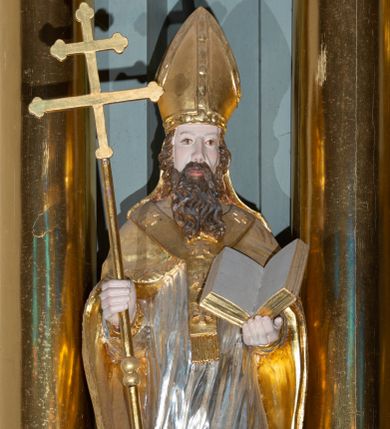 Zdjęcie nr 1: Rzeźba pełnoplastyczna, przyścienna, z tyłu drążona, ustawiona na niskim, prostopadłościennym postumencie. Święty ukazany en face, w kontrapoście, obie ręce ugięte w łokciach, wyciągnięte przed siebie, w lewej trzyma otwartą księgę, w prawej drzewce u dołu zakończone piórem wiosła, zwieńczone krzyżem arcybiskupim. Twarz pociągła, o płytko rzeźbionych rysach i dużym, spiczastym nosie, okolona długą brodą rozdzieloną na dwie części oraz krótkimi falowanymi włosami. Ubrany w długą albę, komżę z koronką na brzegach, kapę spiętą na piersi i opadającą na ramiona i plecy, z fragmentem tkaniny zawiniętym przy prawym boku; na piersi paliusz, na głowie infuła. Figura polichromowana naturalistycznie w odsłoniętych partiach ciała, szaty i atrybuty złocone, komża srebrzona.