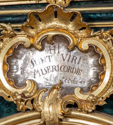 Zdjęcie nr 79: Nastawa ołtarzowa architektoniczna, jednokondygnacyjna ze zwieńczeniem, jednoosiowa z bramkami. Ołtarz prostopadłościenny, murowany, w drewnianej obudowie, z drewnianą mensą. Antepedium ujęte profilowaną ramą; w polu malowana płycina w formie leżącego prostokąta o zaokrąglonych narożach, wypełniona gałązkami różanymi, z akantowo-regencyjnym kartuszem. W narożach antepedium malowane ptaki siedzące na gałązkach. W polu kartusza ukazana scena męczeństwa św. Szymona i św. Judy Tadeusza, na tle pejzażu. Tabernakulum metalowe, prostopadłościenne, flankowane pilastrami w formie spływów wolutowych zdobionych gałązkami róży, zamknięte od góry profilowanym gzymsem. Drzwiczki w kształcie stojącego prostokąta zamkniętego łukiem odcinkowym nadwieszonym; w polu przedstawienie kielicha z hostią. Tabernakulum wprawione w wysoki cokół, na którym stoi tron wystawienia w formie obrotowej niszy, o wypukłej ściance frontowej i ustawionymi pod kątem ściankami bocznymi. Nisza flankowana parą kolumienek i spływów wolutowych dźwigających belkowanie oraz fragmenty przerwanego, półkolistego przyczółka. Na belkowaniu szczyt z rocaillem, z napisem w polu: „ECCE / TABERNACULUM / DEI”. Nastawa ustawiona na dwustrefowym cokole, z wyodrębnionymi czterema prostopadłościanami, w górnej części zdobionym płycinami. Retabulum ujęte parami pilastrów i filarów o kanelowanych trzonach oraz stojącymi przed nimi w układzie schodkowym kolumnami o kapitelach kompozytowych. Podpory dźwigają pełne belkowanie, wyłamane na ich osi, z przerwanym architrawem nad polem środkowym. Na belkowaniu fragmenty falistego, przerwanego przyczółka z figurami aniołów z arma Christi. W polu środkowym wnęka w formie stojącego prostokąta zamkniętego łukiem odcinkowym nadwieszonym, ujęta profilowaną ramą, z obrazem Najświętszej Marii Panny Niepokalanie Poczętej. Nad nią, w polu belkowania rocaille'owy kartusz z napisem „HI / SUNT VIRI / MISERICORDIAE / [...]ccdis 44”. Zwieńczenie w formie zbliżonej do stojącego prostokąta, ujętego pilastrami przechodzącymi w spływy wolutowe, zamkniętego dwoma fragmentami wolutowego gzymsu. Całość zakończona szczytem utworzonym z muszli i zwisów kwiatowych, flankowanym przez dwa wazony. Zwieńczenie ujęte liśćmi palmowymi. W polu obraz z grupą Ukrzyżowania w formie stojącego prostokąta, w profilowanej ramie, dekorowanej rocaille'm. Nastawa flankowana bramkami zwieńczonymi konsolami, na których stoją figury Marii i św. Jana Ewangelisty. Przejścia w formie stojącego prostokąta o wklęsłych górnych narożach, z drzwiczkami z malowanymi przedstawieniami św. Piotra i św. Pawła. Struktura polichromowana na sposób imitujący marmoryzację w kolorze zielono-niebieskim, z laserunkiem, kolumny, ornamentyka i profile złocone, detal srebrzony.