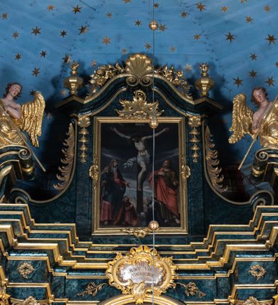 Zdjęcie nr 6: Obraz w formie stojącego prostokąta z przedstawieniem sceny Ukrzyżowania z Matką Boską i św. Janem Ewangelistą oraz dwójką fundatorów. W centrum kompozycji ukazany krzyż z postacią umarłego Chrystusa przybitą trzema gwoździami. Ciało w lekkim zwisie, o szeroko rozłożonych ramionach, z głową opadającą na prawy bark oraz ugiętymi nogami skierowanymi w lewo. Twarz ma szczupłą, o dużych, zamkniętych oczach, prostym nosie i małych ustach, okoloną krótką, ciemną brodą i włosami. Na głowie ma koronę cierniową. Ciało Chrystusa jest silnie umięśnione, z podkreślonymi żebrami i talią klatki piersiowej, z zaznaczonymi śladami męki. Przez biodra przewiązane krótkie, białe perizonium ze zwisem tkaniny rozwianym przy prawym boku. U dołu krzyża leży czaszka i piszczel. Pionowa belka zwieńczona jest tabliczką z napisem „I.N.R.I.”. 
Po prawej stronie krzyża stoi Matka Boska, zwrócona w trzech czwartych w lewo, w kontrapoście, z pochyloną głową i rękami rozłożonymi na boki. Twarz ma kwadratową, o dużych, załzawionych oczach, prostym nosie i pełnych ustach. Ubrana jest w jasnoróżową suknię, przepasaną w talii oraz ciemnoniebieski płaszcz zarzucony na ramiona i zawiązany z przodu postaci. Wokół twarzy ma białą podwikę, na głowie jasnobrązowy welon. 
Po lewej stronie krzyża św. Jan zwrócony w trzech czwartych w prawo, w kontrapoście, z uniesioną głową. Prawą rękę kładzie na piersi, lewą wyciąga w bok. Twarz ma szczupłą, o dużych, załzawionych oczach, prostym nosie i pełnych ustach, okoloną jasnymi włosami. Ubrany jest w zieloną tunikę z kołnierzykiem, przepasaną w talii oraz zawiązany diagonalnie przez prawe ramię jasnoczerwony płaszcz. 
U dołu krzyża klęczą dwie, niewielkie postacie mężczyzny i kobiety, zwrócone do wewnątrz, z uniesionymi głowami i rękami złożonymi w geście modlitwy. Po prawej stronie krzyża mężczyzna z twarzą o cechach portretowych, okoloną brodą z wąsami, z czupryną na głowie. Ubrany w jasnoróżowy żupan z pasem oraz delię, u jego nóg leży kołpak z futrzanym otokiem. Przed nim tarcza ze stylizowanymi labrami i klejnotem, w której godło herbu Junosza. Po lewej kobieta z twarzą o cechach portretowych. Ubrana w czarną suknię z furtygałem, czarny płaszcz z pelerynką. Na głowie ma biały czepiec z białą podwiką i kołpakiem. Przed nią tarcza z godłem herbu Nowina. Scena ukazana na tle pejzażu miejskiego widocznego w oddali, z ciemnym niebem w górnej części obrazu. 

