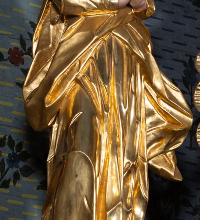 Zdjęcie nr 7: Rzeźba drążona, ustawiona na niskiej podstawie na planie prostokąta. Matka Boska zwrócona w trzech czwartych w lewo, w kontrapoście, lekko odchylona, z głową skierowaną do góry. Dłonie ma splecione z przodu. Twarz ma owalną, z wyraźnie zaznaczoną brodą, o małych, skierowanych w górę oczach, z głęboko rzeźbioną górną powieką, dużym nosie i pełnych ustach z szeroką górną wargą. Na głowie ma złocony welon. Ubrana jest w złoconą tunikę z długimi rękawami oraz złocony płaszcz otulający postać w pasie, rozwiany po bokach. Na stopach ma złocone sandały. Polichromia ciała naturalistyczna. 
