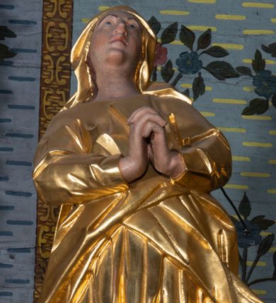 Zdjęcie nr 1: Rzeźba drążona, ustawiona na niskiej podstawie na planie prostokąta. Matka Boska zwrócona w trzech czwartych w lewo, w kontrapoście, lekko odchylona, z głową skierowaną do góry. Dłonie ma splecione z przodu. Twarz ma owalną, z wyraźnie zaznaczoną brodą, o małych, skierowanych w górę oczach, z głęboko rzeźbioną górną powieką, dużym nosie i pełnych ustach z szeroką górną wargą. Na głowie ma złocony welon. Ubrana jest w złoconą tunikę z długimi rękawami oraz złocony płaszcz otulający postać w pasie, rozwiany po bokach. Na stopach ma złocone sandały. Polichromia ciała naturalistyczna. 
