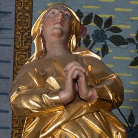 Zdjęcie nr 1: Rzeźba drążona, ustawiona na niskiej podstawie na planie prostokąta. Matka Boska zwrócona w trzech czwartych w lewo, w kontrapoście, lekko odchylona, z głową skierowaną do góry. Dłonie ma splecione z przodu. Twarz ma owalną, z wyraźnie zaznaczoną brodą, o małych, skierowanych w górę oczach, z głęboko rzeźbioną górną powieką, dużym nosie i pełnych ustach z szeroką górną wargą. Na głowie ma złocony welon. Ubrana jest w złoconą tunikę z długimi rękawami oraz złocony płaszcz otulający postać w pasie, rozwiany po bokach. Na stopach ma złocone sandały. Polichromia ciała naturalistyczna. 
