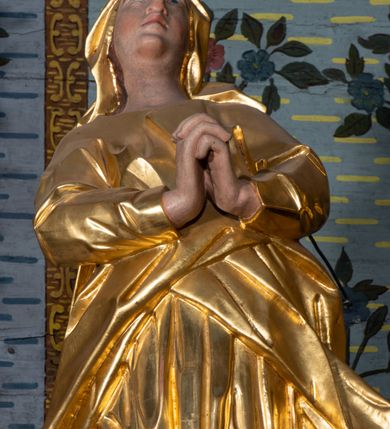 Zdjęcie nr 9: Rzeźba drążona, ustawiona na niskiej podstawie na planie prostokąta. Matka Boska zwrócona w trzech czwartych w lewo, w kontrapoście, lekko odchylona, z głową skierowaną do góry. Dłonie ma splecione z przodu. Twarz ma owalną, z wyraźnie zaznaczoną brodą, o małych, skierowanych w górę oczach, z głęboko rzeźbioną górną powieką, dużym nosie i pełnych ustach z szeroką górną wargą. Na głowie ma złocony welon. Ubrana jest w złoconą tunikę z długimi rękawami oraz złocony płaszcz otulający postać w pasie, rozwiany po bokach. Na stopach ma złocone sandały. Polichromia ciała naturalistyczna. 
