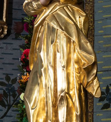 Zdjęcie nr 2: Rzeźba drążona, ustawiona na niskiej podstawie na planie prostokąta. Matka Boska zwrócona w trzech czwartych w lewo, w kontrapoście, lekko odchylona, z głową skierowaną do góry. Dłonie ma splecione z przodu. Twarz ma owalną, z wyraźnie zaznaczoną brodą, o małych, skierowanych w górę oczach, z głęboko rzeźbioną górną powieką, dużym nosie i pełnych ustach z szeroką górną wargą. Na głowie ma złocony welon. Ubrana jest w złoconą tunikę z długimi rękawami oraz złocony płaszcz otulający postać w pasie, rozwiany po bokach. Na stopach ma złocone sandały. Polichromia ciała naturalistyczna. 
