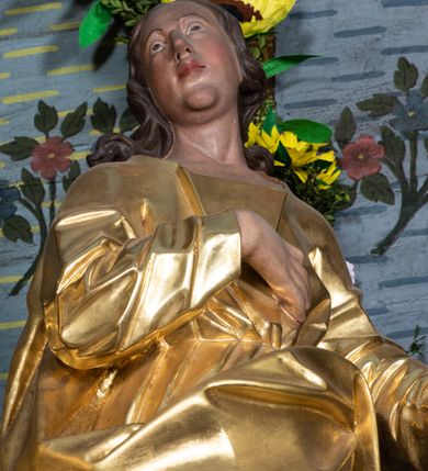 Zdjęcie nr 9: Rzeźba drążona, ustawiona na niskiej podstawie na planie prostokąta. Święty zwrócony w trzech czwartych w prawo, w delikatnym kontrapoście, z lekko uniesioną głową, z lewą ręką wyciągniętą w bok, prawą złożoną na piersi. Twarz ma owalną, z wyraźnie zaznaczoną brodą, o małych oczach zwróconych w górę, z głęboko rzeźbioną górną powieką, dużym nosem i pełnymi ustami z szeroką górną wargą. Włosy ma ciemnobrązowe, silnie kręcone, opadające na ramiona. Ubrany jest w złoconą tunikę z długimi rękawami, przepasaną w talii oraz złocony płaszcz zawiązany z przodu postaci, na lewym biodrze. Na stopach ma złocone sandały. Polichromia naturalistyczna w partiach ciała. 