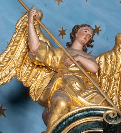 Zdjęcie nr 6: Rzeźby drążone z tyłu, klęczące na fragmentach przerwanego przyczółka, zwrócone do wewnątrz, pochylone. Lewy anioł w uniesionej lewej ręce trzyma laskę zakończoną gąbką, prawą kładzie na piersi. Prawy anioł obiema rękami trzyma włócznię. Twarze mają pociągłe, z wyodrębnionymi brodami, o małych oczach z głęboko rzeźbionymi górnymi powiekami, krótkimi nosami i pełnymi ustami, okolone kręconymi, brązowymi włosami, sięgającymi ramion. Anioły ubrane są w złocone tuniki z jednym rękawem, odsłaniające połowę torsu i drugie ramię, przepasane w talii. Skrzydła mają duże i złożone po bokach. Polichromia ciał naturalistyczna. 

