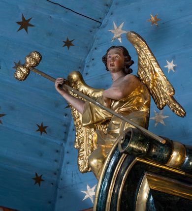 Zdjęcie nr 5: Rzeźby drążone z tyłu, klęczące na fragmentach przerwanego przyczółka, zwrócone do wewnątrz, pochylone. Lewy anioł w uniesionej lewej ręce trzyma laskę zakończoną gąbką, prawą kładzie na piersi. Prawy anioł obiema rękami trzyma włócznię. Twarze mają pociągłe, z wyodrębnionymi brodami, o małych oczach z głęboko rzeźbionymi górnymi powiekami, krótkimi nosami i pełnymi ustami, okolone kręconymi, brązowymi włosami, sięgającymi ramion. Anioły ubrane są w złocone tuniki z jednym rękawem, odsłaniające połowę torsu i drugie ramię, przepasane w talii. Skrzydła mają duże i złożone po bokach. Polichromia ciał naturalistyczna. 

