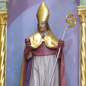 Zdjęcie nr 1: Rzeźba pełnoplastyczna, polichromowana, ustawiona na niewielkiej, prostopadłościennej podstawie. Święty ukazany całopostaciowo, frontalnie, w lekkim kontrapoście, z głową zwróconą w prawo oraz obiema rękami ugiętymi w łokciu i wyciągniętymi przed siebie. W lewym ręku trzyma złocony pastorał z akantową krzywaśnią i srebrzonym drzewcem. Święty przedstawiony jako młody mężczyzna o lekko falowanych, ciemnych włosach i twarzy pozbawionej zarostu. Twarz szczupła, owalna z niewielkim, prostym nosem oraz małych, nieznacznie rozchylonych ustach i zarumienionych policzkach. Postać ubrana w szaty biskupie: ciemnoczerwoną sutannę, białą komżę wykończoną koronką, ciemnoczerwony mucet oraz złocony, obszerny płaszcz z czerwoną podszewką, spięty pod szyją. Na głowie złocona infuła biskupia dekorowana równoramiennym krzyżem. Na rękach ciemnoczerwone rękawiczki.