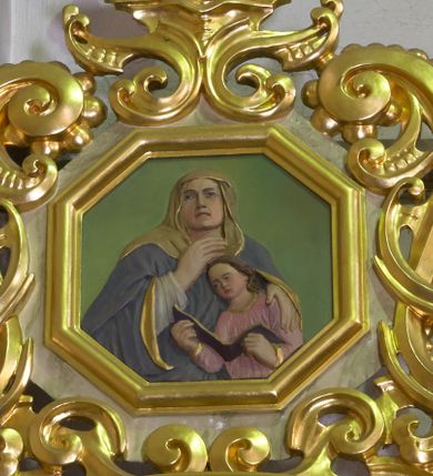 Zdjęcie nr 1: Płaskorzeźba w kształcie ośmioboku ujęta profilowaną, złoconą ramką oraz ażurowym obramieniem utworzonym z przeciwstawnie ułożonych wolut wzbogaconych ornamentem małżowinowo-chrząstkowym. W zwieńczeniu złocony krzyż o trójlistnie zakończonych ramionach ustawiony na niewielkim postumencie wspartym na strukturze z ornamentu małżowinowo-chrząstkowego. W centralnym polu płaskorzeźbione przedstawienie św. Anny w towarzystwie młodzieńczej Marii. Święta ukazana w popiersiu, frontalnie, z głową uniesioną nieznacznie ku górze. Anna została przedstawiona jako dojrzała kobieta ubrana w białą szatę, obszerny, ciemnogranatowy płaszcz ze złotą podszewką oraz żółtą chustę zarzuconą na głowę. Swoją prawą dłoń przyciska do piersi, lewą obejmuje przedstawioną w jasnoróżowej szacie Marię, która czyta z otwartej księgi. Tło jednolicie jasnozielone, postaci polichromowane; partie ornamentu oraz detale szat złocone.

