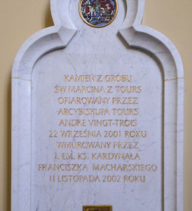 Zdjęcie nr 1: Kamienna tablica pamiątkowa w kształcie stojącego prostokąta  zamkniętego ostrym łukiem trójlistnym  w profilowanej ramie. W górnej części medalion z emaliowanym przedstawieniem  św. Marcina na koniu dzielącego się płaszczem z żebrakiem. W części centralnej złocona inskrypcja „KAMIEŃ Z GROBU / ŚW[IĘTEGO] MARCINA Z TOURS / OFIAROWANY PRZEZ / ARCYBISKUPA TOURS / ANDRE VINGT-TROIS / 22 WRZEŚNIA 2001 ROKU / WMUROWANY PRZEZ / J(EGO) EM(INENCJĘ) KS(IĘDZA) KARDYNAŁA / FRANCISZKA MACHARSKIEGO / 11 LISTOPADA 2002 ROKU”. Poniżej inskrypcji mała kwadratowa, złocona, przeszklona nisza zawierająca kamień z grobu św. Marcina z Tours.
