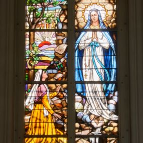 Zdjęcie nr 1: Witraż w drugim przęśle od zachodu, w oknie południowym. Kompozycja w kształcie stojącego prostokąta zamkniętego łukiem ostrym, podzielona pionową laską na dwie części, u góry z maswerkiem o motywie czteroliścia. Witraż ukazuje scenę objawienia się Matki Boskiej w Lourdes. W lewej części Matka Boska stojąca w grocie, w białej sukni, błękitnym płaszczu, przewiązana w pasie szarfą. Na prawej ręce ma zawieszony różaniec, wokół głowy dziesięć gwiazd. Spod stóp Marii wypływa źródło. W prawej części klęcząca św. Bernadeta ubrana w pomarańczową suknię, fioletową chustkę, na głowie biały welon. W tle pejzaż z wschodzącym słońcem. W górnej części dekoracja ornamentalna w postaci geometrycznych rozet. W polu czteroliścia godło z koroną, jabłkiem oraz skrzyżowanym berłem i mieczem. W dolnej części róże tworzące dekoracyjny pas. W prawym dolnym narożu napis: RYS. JAN MIKOŁAJSKI / ODREMONTOWALI / BRACIA PACZKA. U dołu prostokątne, białe pole z napisem: FUNDAVIT HEDVIGIS ORTYŃSKA EMERITA / MAGISTRA SCHOLAE / A.D. 1939 CURA PAROCHI JACOBI MORAJKA. Bogaty modelunek światłocieniowy, żywa kolorystyka.