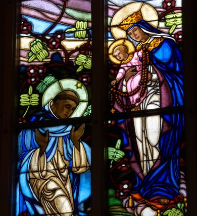 Zdjęcie nr 1: Witraż w prezbiterium w oknie południowym, kształcie stojącego prostokąta zamkniętego łukiem ostrym. Kompozycja podzielona w pionową laską na dwie części, u góry z maswerkiem o motywie czteroliścia. W centrum przestawienie stojącej Marii w białej szacie i niebieskim płaszczu, z koroną na głowie, trzymającej na ramionach Chrystusa, który przekazuje różaniec klęczącemu po lewej stronie św. Dominikowi. Święty ubrany w biały habit oraz błękitny płaszcz. U jego stop leży księga oraz płonąca pochodnia. W tle pejzaż oraz bogata dekoracja roślinna. W górnej części, pod maswerkiem hierogram Chrystusa i splecione litery imienia Maria w obramieniach w kształcie mandorli. W polu czteroliścia przedstawienie kielicha z hostią, po bokach z kiścią winogron i kłosem pszenicy. Dolną część kompozycji wypełnia dekoracja roślinna, w którą wplecione są dwa medaliony, po lewej z sercem Chrystusa (płonące serce, ujęte cierniową koroną, zwieńczone krzyżem), po prawej serce Marii (płonące serce oplecione wieńcem z białych róż, przebite mieczem) – oba serca ujęte glorią promienistą.
