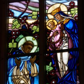 Zdjęcie nr 1: Witraż w prezbiterium w oknie południowym, kształcie stojącego prostokąta zamkniętego łukiem ostrym. Kompozycja podzielona w pionową laską na dwie części, u góry z maswerkiem o motywie czteroliścia. W centrum przestawienie stojącej Marii w białej szacie i niebieskim płaszczu, z koroną na głowie, trzymającej na ramionach Chrystusa, który przekazuje różaniec klęczącemu po lewej stronie św. Dominikowi. Święty ubrany w biały habit oraz błękitny płaszcz. U jego stop leży księga oraz płonąca pochodnia. W tle pejzaż oraz bogata dekoracja roślinna. W górnej części, pod maswerkiem hierogram Chrystusa i splecione litery imienia Maria w obramieniach w kształcie mandorli. W polu czteroliścia przedstawienie kielicha z hostią, po bokach z kiścią winogron i kłosem pszenicy. Dolną część kompozycji wypełnia dekoracja roślinna, w którą wplecione są dwa medaliony, po lewej z sercem Chrystusa (płonące serce, ujęte cierniową koroną, zwieńczone krzyżem), po prawej serce Marii (płonące serce oplecione wieńcem z białych róż, przebite mieczem) – oba serca ujęte glorią promienistą.
