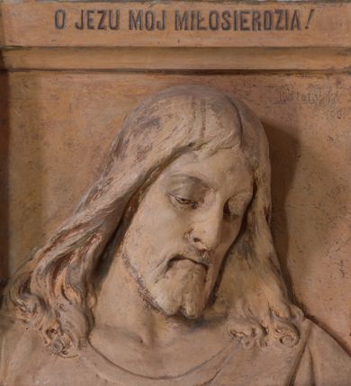 Zdjęcie nr 1: Płaskorzeźba w kształcie stojącego prostokąta z przedstawieniem w popiersiu Chrystusa Miłosiernego ujętego od dołu i od góry poziomymi listwami. Chrystus ukazany frontalnie, z głową lekko pochyloną i przechyloną na lewy bark. Twarz pociągła, z wydatnym nosem, wpółprzymkniętym oczami i wzrokiem skierowanym w dół; z wąsami i krótką, rozdwojoną na końcu brodą. Włosy długie, pofalowane, opadające na ramiona okryte cienką suknią z półkolistym wycięciem pod szyją. U góry na profilowanej belce inskrypcja majuskułą po polsku: O JEZU, z krót MÓJ MIŁOSIERDZIA!  Na dolnej listwie inskrypcja: Fund. Parochus Jacobus M(orajka) 1933. Całość pomalowana w kolorze jasnego beżu z przetarciami warstwy malarskiej.
