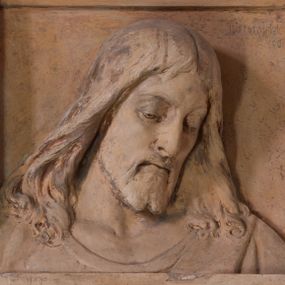 Zdjęcie nr 1: Płaskorzeźba w kształcie stojącego prostokąta z przedstawieniem w popiersiu Chrystusa Miłosiernego ujętego od dołu i od góry poziomymi listwami. Chrystus ukazany frontalnie, z głową lekko pochyloną i przechyloną na lewy bark. Twarz pociągła, z wydatnym nosem, wpółprzymkniętym oczami i wzrokiem skierowanym w dół; z wąsami i krótką, rozdwojoną na końcu brodą. Włosy długie, pofalowane, opadające na ramiona okryte cienką suknią z półkolistym wycięciem pod szyją. U góry na profilowanej belce inskrypcja majuskułą po polsku: O JEZU, z krót MÓJ MIŁOSIERDZIA!  Na dolnej listwie inskrypcja: Fund. Parochus Jacobus M(orajka) 1933. Całość pomalowana w kolorze jasnego beżu z przetarciami warstwy malarskiej.