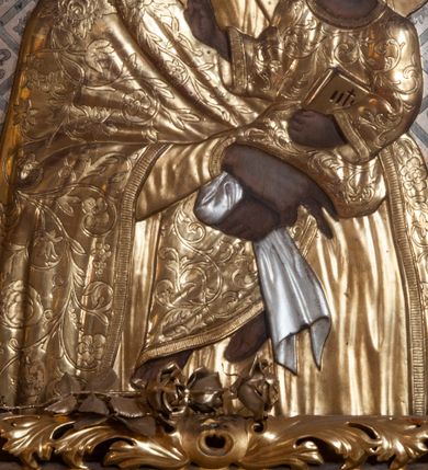 Zdjęcie nr 4: Matka Boska z Dzieciątkiem w typie Śnieżnej, ujęta w półfigurze, en face, z lekkim skrętem głowy w prawo, ku podtrzymywanemu na lewym przedramieniu – Dzieciątku. Twarz Matki Boskiej pociągła o regularnych rysach; cienkich brwiach, długim nosie, małych ustach, okolona welonem odwijającym się fałdami od twarzy, opadającym szerokim płaszczem na ramiona. Suknia marszczona w gęste, drobne, pionowe fałdy. Ręce splecione na nóżkach Dzieciątka, z palcami prawej dłoni skierowanymi w dół. Dzieciątko o główce ujętej w ¾, zwróconej ku Marii, okolonej krótkimi, kręconymi włosami. Prawa rękę wznosi w geście błogosławienia, lewa podtrzymuje księgę. Odziane w długą sukienkę, o szerokich rękawach, spod której widoczne bose stopy. Sukienki drewniane, złocone. Płaszcz M. Boskiej i sukienka Dzieciątka pokryte ornamentem o motywie roślinnym, na głowach półplastyczne korony, wokół – nimby koliste, zdobione promieniami. Tło w formie ukośnej kratownicy, wypełnionej lilijkami i geometrycznymi rozetkami. Rama ażurowa, z motywami liści akantu i spinaczami w narożach. 