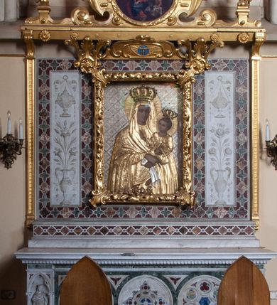 Zdjęcie nr 5: Matka Boska z Dzieciątkiem w typie Śnieżnej, ujęta w półfigurze, en face, z lekkim skrętem głowy w prawo, ku podtrzymywanemu na lewym przedramieniu – Dzieciątku. Twarz Matki Boskiej pociągła o regularnych rysach; cienkich brwiach, długim nosie, małych ustach, okolona welonem odwijającym się fałdami od twarzy, opadającym szerokim płaszczem na ramiona. Suknia marszczona w gęste, drobne, pionowe fałdy. Ręce splecione na nóżkach Dzieciątka, z palcami prawej dłoni skierowanymi w dół. Dzieciątko o główce ujętej w ¾, zwróconej ku Marii, okolonej krótkimi, kręconymi włosami. Prawa rękę wznosi w geście błogosławienia, lewa podtrzymuje księgę. Odziane w długą sukienkę, o szerokich rękawach, spod której widoczne bose stopy. Sukienki drewniane, złocone. Płaszcz M. Boskiej i sukienka Dzieciątka pokryte ornamentem o motywie roślinnym, na głowach półplastyczne korony, wokół – nimby koliste, zdobione promieniami. Tło w formie ukośnej kratownicy, wypełnionej lilijkami i geometrycznymi rozetkami. Rama ażurowa, z motywami liści akantu i spinaczami w narożach. 