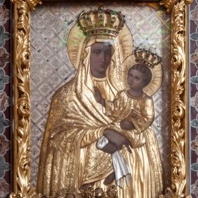 Zdjęcie nr 1: Matka Boska z Dzieciątkiem w typie Śnieżnej, ujęta w półfigurze, en face, z lekkim skrętem głowy w prawo, ku podtrzymywanemu na lewym przedramieniu – Dzieciątku. Twarz Matki Boskiej pociągła o regularnych rysach; cienkich brwiach, długim nosie, małych ustach, okolona welonem odwijającym się fałdami od twarzy, opadającym szerokim płaszczem na ramiona. Suknia marszczona w gęste, drobne, pionowe fałdy. Ręce splecione na nóżkach Dzieciątka, z palcami prawej dłoni skierowanymi w dół. Dzieciątko o główce ujętej w ¾, zwróconej ku Marii, okolonej krótkimi, kręconymi włosami. Prawa rękę wznosi w geście błogosławienia, lewa podtrzymuje księgę. Odziane w długą sukienkę, o szerokich rękawach, spod której widoczne bose stopy. Sukienki drewniane, złocone. Płaszcz M. Boskiej i sukienka Dzieciątka pokryte ornamentem o motywie roślinnym, na głowach półplastyczne korony, wokół – nimby koliste, zdobione promieniami. Tło w formie ukośnej kratownicy, wypełnionej lilijkami i geometrycznymi rozetkami. Rama ażurowa, z motywami liści akantu i spinaczami w narożach. 