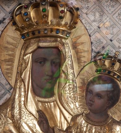 Zdjęcie nr 2: Matka Boska z Dzieciątkiem w typie Śnieżnej, ujęta w półfigurze, en face, z lekkim skrętem głowy w prawo, ku podtrzymywanemu na lewym przedramieniu – Dzieciątku. Twarz Matki Boskiej pociągła o regularnych rysach; cienkich brwiach, długim nosie, małych ustach, okolona welonem odwijającym się fałdami od twarzy, opadającym szerokim płaszczem na ramiona. Suknia marszczona w gęste, drobne, pionowe fałdy. Ręce splecione na nóżkach Dzieciątka, z palcami prawej dłoni skierowanymi w dół. Dzieciątko o główce ujętej w ¾, zwróconej ku Marii, okolonej krótkimi, kręconymi włosami. Prawa rękę wznosi w geście błogosławienia, lewa podtrzymuje księgę. Odziane w długą sukienkę, o szerokich rękawach, spod której widoczne bose stopy. Sukienki drewniane, złocone. Płaszcz M. Boskiej i sukienka Dzieciątka pokryte ornamentem o motywie roślinnym, na głowach półplastyczne korony, wokół – nimby koliste, zdobione promieniami. Tło w formie ukośnej kratownicy, wypełnionej lilijkami i geometrycznymi rozetkami. Rama ażurowa, z motywami liści akantu i spinaczami w narożach. 