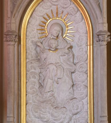 Zdjęcie nr 1: Płaskorzeźba w kształcie prostokąta zamkniętego ostrołukowo. Przedstawienie tronującej na obłokach Matki Boskiej z Dzieciątkiem. Jezus, ubrany w długą sukienkę z długimi rękawami, siedzi na kolanach Marii z uniesionymi rozłożonymi na boki ramionami. Wokół głowy Marii złocony nimb w kształcie okręgu z promieniami wokół.