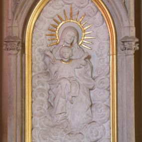 Zdjęcie nr 1: Płaskorzeźba w kształcie prostokąta zamkniętego ostrołukowo. Przedstawienie tronującej na obłokach Matki Boskiej z Dzieciątkiem. Jezus, ubrany w długą sukienkę z długimi rękawami, siedzi na kolanach Marii z uniesionymi rozłożonymi na boki ramionami. Wokół głowy Marii złocony nimb w kształcie okręgu z promieniami wokół.