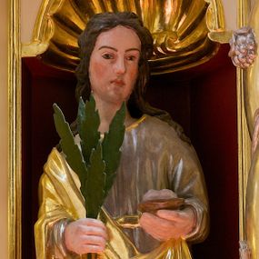 Zdjęcie nr 1: Figura pełna przedstawiająca nieokreśloną świętą męczennicę. Święta stoi frontalnie, spoglądając lekko w lewą stronę. Ma podłużną twarz, małe oczy, wąski nos, niewielkie usta i długie, ciemne włosy spływające falami na ramiona i plecy. W prawej ręce trzyma palmę męczeńską, w lewej trudny do określenia przedmiot umieszczony na podstawce. Ubrana jest w przepasaną, srebrną suknię wykończoną przy dekolcie złotą taśmą oraz złoty płaszcz, który przechodzi przez prawą rękę i jest podczepiony na lewym biodrze. 