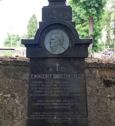 Zdjęcie nr 1: Nagrobek wolnostojący w kształcie krzyża ustawionego na dwustrefowym, wysokim cokole ze zwieńczeniem zamkniętym łukiem nadwieszonym, w którego podłuczu znajduje się płaskorzeźbiony profil zmarłego. Na przecięciu ramion krzyża ustawionego na profilowanym cokole widnieje chrystogram. Na czołowym boku cokołu wyryto napis „A. †  Ω. / X(IĄDZ) WINCENTY SMOCZYŃSKI S(WIĘTEJ) T(EOLOGII) D(OKTOR) / PROBOSZCZ PARAFII TENCZYŃSKIEJ A NASTĘPNIE / PREPOZYT KOLLEGIATY ŚW(IĘTEGO) FLORYANA W KRAKOWIE, / TAJNY PODKOMORZY PAPIESKI, KANONIK H(ONOROWY) KALISKI / CZŁONEK AKADEMII RZYMSKIEJ „DEGLI ARCADI” / UR(ODZONY) 7. LISTOPADA 1842. ZM(ARŁY) 10. SIERPNIA 1903. W KRAKOWIE. / 
SZEDŁ PO CIERNISTEJ DRODZE NIOSĄC KRZYŻ / ZA CHRYSTUSEM PANEM. NIECHŻE GO PAN NAJWYŻSZY POLICZY / DO LICZBY BŁOGOSŁAWIONYCH. / 
ZACNEMU I UKOCHANEMU KAPŁANOWI, PASTERZOWI I BRATU / POMNIK TEN POSTAWIŁ X(IĄDZ) KANONIK IGNACY MECH KAPŁAN Z DYECEZYI LUBELSKIEJ / PRZYJACIEL I TOWARZYSZ CHWIL POWOŁANIA DUCHOWNEGO Z AKADEMII DUCH(OWNEJ) WARSZAWSKIEJ / WRAZ Z SIOSTRAMI ZMARŁEGO. / R(EQUIESCAT) I(N) P(ACE)”.