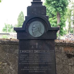 Zdjęcie nr 1: Nagrobek wolnostojący w kształcie krzyża ustawionego na dwustrefowym, wysokim cokole ze zwieńczeniem zamkniętym łukiem nadwieszonym, w którego podłuczu znajduje się płaskorzeźbiony profil zmarłego. Na przecięciu ramion krzyża ustawionego na profilowanym cokole widnieje chrystogram. Na czołowym boku cokołu wyryto napis „A. †  Ω. / X(IĄDZ) WINCENTY SMOCZYŃSKI S(WIĘTEJ) T(EOLOGII) D(OKTOR) / PROBOSZCZ PARAFII TENCZYŃSKIEJ A NASTĘPNIE / PREPOZYT KOLLEGIATY ŚW(IĘTEGO) FLORYANA W KRAKOWIE, / TAJNY PODKOMORZY PAPIESKI, KANONIK H(ONOROWY) KALISKI / CZŁONEK AKADEMII RZYMSKIEJ „DEGLI ARCADI” / UR(ODZONY) 7. LISTOPADA 1842. ZM(ARŁY) 10. SIERPNIA 1903. W KRAKOWIE. / 
SZEDŁ PO CIERNISTEJ DRODZE NIOSĄC KRZYŻ / ZA CHRYSTUSEM PANEM. NIECHŻE GO PAN NAJWYŻSZY POLICZY / DO LICZBY BŁOGOSŁAWIONYCH. / 
ZACNEMU I UKOCHANEMU KAPŁANOWI, PASTERZOWI I BRATU / POMNIK TEN POSTAWIŁ X(IĄDZ) KANONIK IGNACY MECH KAPŁAN Z DYECEZYI LUBELSKIEJ / PRZYJACIEL I TOWARZYSZ CHWIL POWOŁANIA DUCHOWNEGO Z AKADEMII DUCH(OWNEJ) WARSZAWSKIEJ / WRAZ Z SIOSTRAMI ZMARŁEGO. / R(EQUIESCAT) I(N) P(ACE)”.