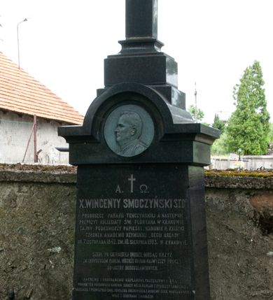 Zdjęcie nr 2: Nagrobek wolnostojący w kształcie krzyża ustawionego na dwustrefowym, wysokim cokole ze zwieńczeniem zamkniętym łukiem nadwieszonym, w którego podłuczu znajduje się płaskorzeźbiony profil zmarłego. Na przecięciu ramion krzyża ustawionego na profilowanym cokole widnieje chrystogram. Na czołowym boku cokołu wyryto napis „A. †  Ω. / X(IĄDZ) WINCENTY SMOCZYŃSKI S(WIĘTEJ) T(EOLOGII) D(OKTOR) / PROBOSZCZ PARAFII TENCZYŃSKIEJ A NASTĘPNIE / PREPOZYT KOLLEGIATY ŚW(IĘTEGO) FLORYANA W KRAKOWIE, / TAJNY PODKOMORZY PAPIESKI, KANONIK H(ONOROWY) KALISKI / CZŁONEK AKADEMII RZYMSKIEJ „DEGLI ARCADI” / UR(ODZONY) 7. LISTOPADA 1842. ZM(ARŁY) 10. SIERPNIA 1903. W KRAKOWIE. / 
SZEDŁ PO CIERNISTEJ DRODZE NIOSĄC KRZYŻ / ZA CHRYSTUSEM PANEM. NIECHŻE GO PAN NAJWYŻSZY POLICZY / DO LICZBY BŁOGOSŁAWIONYCH. / 
ZACNEMU I UKOCHANEMU KAPŁANOWI, PASTERZOWI I BRATU / POMNIK TEN POSTAWIŁ X(IĄDZ) KANONIK IGNACY MECH KAPŁAN Z DYECEZYI LUBELSKIEJ / PRZYJACIEL I TOWARZYSZ CHWIL POWOŁANIA DUCHOWNEGO Z AKADEMII DUCH(OWNEJ) WARSZAWSKIEJ / WRAZ Z SIOSTRAMI ZMARŁEGO. / R(EQUIESCAT) I(N) P(ACE)”.